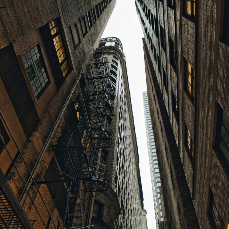 Photograph looking straight up a narrow Chicago alley between old brick buildings with black fire escapes, a sliver of white sky and a distant tower